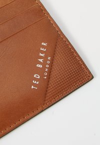 Smeđi kožni novčanik, teksturirana površina, s utorima za kartice i urezanim logotipom "TED BAKER" u srebrnoj boji. Čisto šivanje, trokutasti detalj na uglovima.