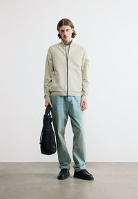 Νέος άνδρας στέκεται κρατώντας μαύρη τσάντα, φοράει μπεζ μπουφάν με φερμουάρ, ανοιχτό μπλε τζιν και μαύρα παπούτσια, σε απλό λευκό φόντο.