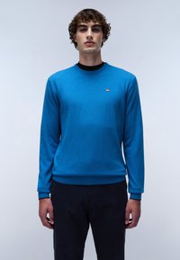 Maglione lavorato a maglia blu con scollo rotondo, caratterizzato da un piccolo logo sul lato sinistro del petto. Indossato con pantaloni scuri, che mettono in risalto un design semplice.