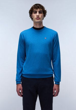 Suéter azul de punto con cuello redondo, que presenta un pequeño logo en el lado izquierdo del pecho. Combinado con pantalones oscuros, que exhiben un diseño sencillo.