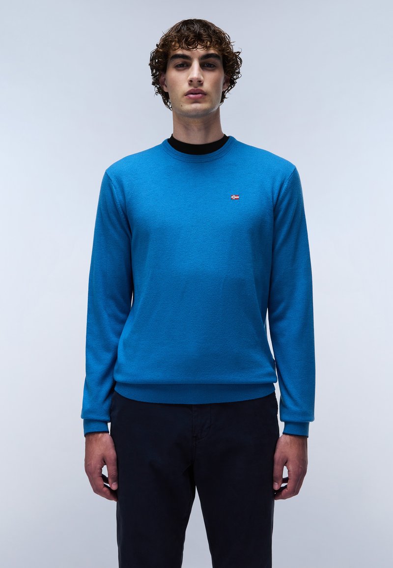 Maglione lavorato a maglia blu con scollo rotondo, caratterizzato da un piccolo logo sul lato sinistro del petto. Indossato con pantaloni scuri, che mettono in risalto un design semplice.