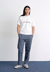 Camiseta blanca de algodón con el logo de "New Balance", pantalones ligeros grises y zapatillas deportivas plateadas. El modelo está de pie en un fondo neutro.