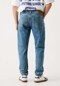 Jeans de mezclilla azules claros con un corte holgado, que cuentan con dos bolsillos traseros y un desgaste sutil. Llevados con zapatillas blancas y una camiseta gráfica.