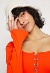 T-shirt laranja canelado de manga longa com decote em forma de barco e fechos decorativos prateados. Um chapéu de balde branco felpudo é usado na cabeça.