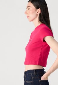 Žena stojí v profilu, oblečená v priliehavom ružovom crop top a tmavých džínoch, s rukou opretou na bok, predvádzajúc moderný štýl.