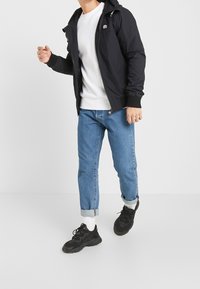 Svart huva jacka med dragkedja, vit t-shirt, ljusblå jeans, uppvikt ärm och svarta sneakers med vita detaljer. Enkelt design och avslappnad passform.