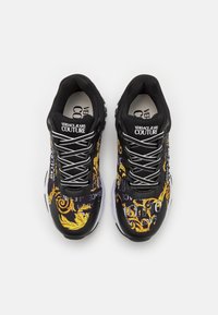 Baskets noires avec une finition brillante, présentant des motifs graphiques jaunes et blancs, des accents de logo et une semelle amortissante.