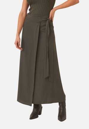 Mujer con falda tipo pareo verde oliva con lazo lateral y botines negros, posando frente a un fondo blanco.