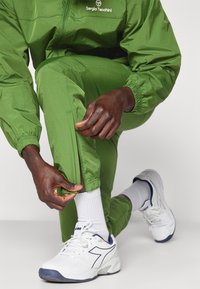 Zaļš treniņtērps ar rāvējslēdzēju potītēs, izgatavots no viegla materiāla. Baltas sneakers ar tumši zilām akcentiem un redzamām ribotām baltām zeķēm.