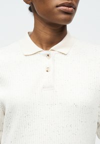 Κοντινό πλάνο ενός ατόμου που φοράει λευκό ριμπ πικέ πόλο με κρεμ γιακά και δύο κουμπιά κλειστά στο λαιμό.