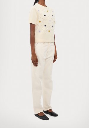 Osoba ubrana w kremowy, krótki sweter z teksturowanymi kropkami i małymi kwiatami, kremowe szerokie spodnie oraz czarne baletki.