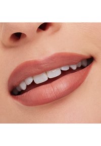 Laboratorio de labios suaves y llenos en un tono mate nude, con dientes blancos visibles. La piel presenta un tono y textura naturales, realzando el aspecto general.