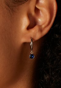 Zilveren oorring met een blauwe edelsteenaccent, met een gepolijste afwerking en een sferische vorm, die net onder de oorschelp hangt.