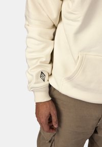 Felpa color crema con polsini a coste, caratterizzata da un logo geometrico nero sulla manica. Tessuto morbido, dettagli della tasca frontale, vestibilità rilassata.