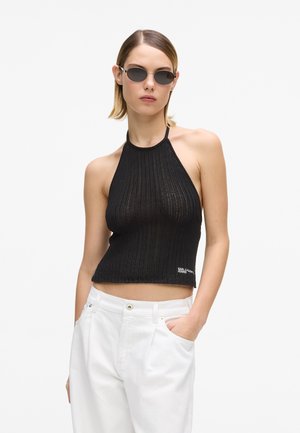 Črna pletena halter majica z rebrasto teksturo, krajšim krojem in logotipom. Kombinirana z belimi hlačami in majhnimi črnimi sončnimi očali.