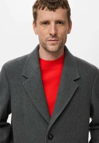 Cappotto grigio in lana con colletto a risvolto e un singolo bottone nero, indossato su un maglione lavorato a maglia rosso brillante. Texture liscia e vestibilità sartoriale.