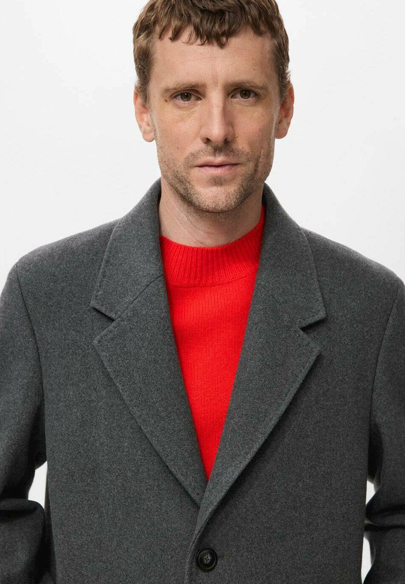 Grå ullkappa med slagkrage och en svart knapp, bärs över en klar röd stickad tröja. Slät textur och skräddarsydd passform.