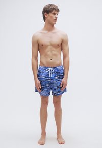 Μπλε μαγιό με σχέδιο κυμάτων, ρυθμιζόμενο λευκό κορδόνι και μήκος μέχρι το μέσο του μηρού. Ομαλή υφή υφάσματος.