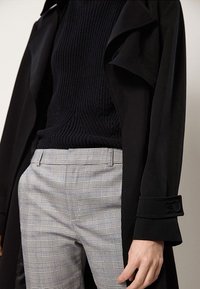 Zwarte trenchcoat met opgevouwen kraag over een marineblauwe geribde trui. Grijs geruite broek met een gecontroleerd patroon en zijzakken.