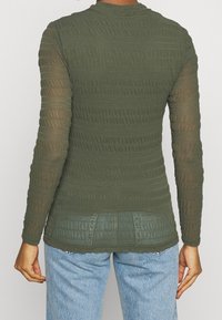 Top a maniche lunghe di colore verde oliva con strisce orizzontali testurizzate e una silhouette aderente. Maniche trasparenti e orlo leggermente accorciato.