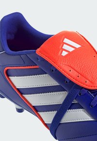 Botín de fútbol azul y naranja con parte superior sintética, que cuenta con tres franjas blancas, una superficie texturizada y una lengüeta acolchada.