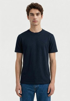 Ung mann med kort brunt hår iført en enkel mørk marineblå T-skjorte med rund hals og blå jeans, stående mot en lys grå bakgrunn.