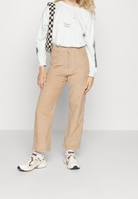 Pantalon en velours côtelé beige à jambes larges, assorti d'une chemise à manches longues bleu clair avec des détails de logo et d'un sac tissé noir et blanc.
