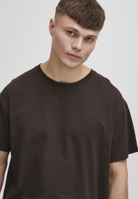 Brązowy bawełniany t-shirt z krótkimi rękawami, z dyskretnym logo w ciemnozielonym kolorze na klatce piersiowej oraz okrągłym dekoltem z przeszytymi detalami.