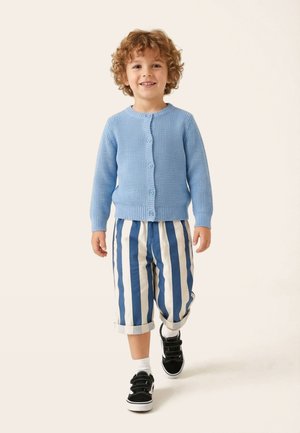 Fiatal, göndör hajú gyermek kék gombos pulóverben és kék-fehér csíkos nadrágban, mosollyal sétál előre.