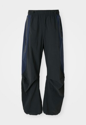 Black outdoor pants with a relaxed fit, featuring elastic waistband, navy side panels, and zippered pockets on both sides.