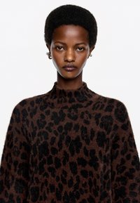 Jersey marrón con un patrón de leopardo negro, cuello alto, textura suave y corte relajado. La modelo lleva pendientes dorados.
