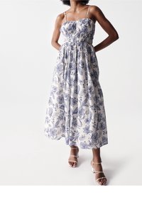 Valkoinen ja laivastonsininen kukkakuosinen maksimekko, jossa on smokkattu yläosa ja ohuet hihnat. Korkea vyötärö ja laskeutuva helma. Yhdistetty valkoisiin remmikkäisiin korkokenkiin.