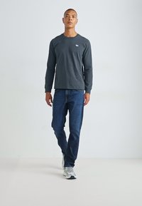 Camisa de manga larga en gris carbón combinada con jeans azules. La camisa presenta un pequeño logo en el pecho. Llevada con zapatillas deportivas de color claro.