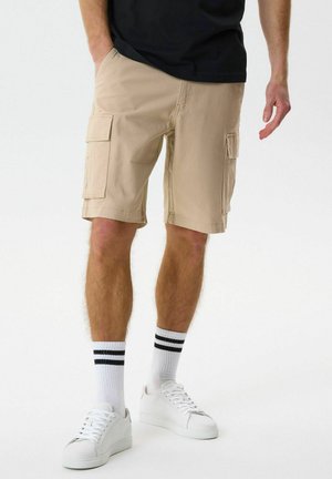 Beige színű terep rövidnadrágot, fehér tornacipőt és fekete csíkos fehér zoknit viselő férfi, aki az egyik kezét a zsebében tartva áll.