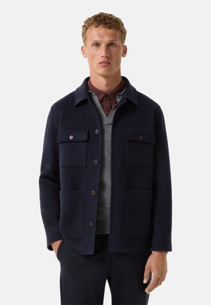 Navy wool blend jacket with a collared design, featuring four front pockets, brown buttons, and a straight cut. Layered over a gray sweater.