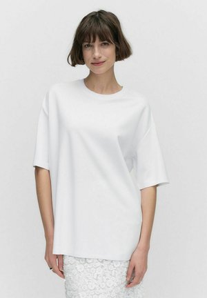 T-shirt blanc surdimensionné en tissu lisse avec des manches courtes et un col rond, associé à une jupe blanche en dentelle texturée.