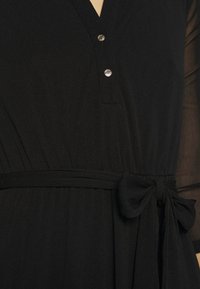 Robe noire avec deux boutons argentés près de l'encolure, manches longues en voile transparent, et un nœud noué en tissu à la taille sur le côté droit.