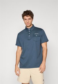 Marineblå polo t-shirt med knapkrave, korte æ sleeves og en brystlomme. Har et lille logo og teksturerede stofdetaljer.