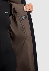 Brun puffjacka med en mjuk textur, med en svart innerfoder. Inkluderar etiketter på både innerfickan och utsidan.