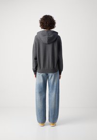 Sudadera gris oscuro con capucha, corte relajado, puños acanalados, combinada con jeans de pierna ancha azul claro y zapatos con suela amarilla. Textura mate, estilo casual.