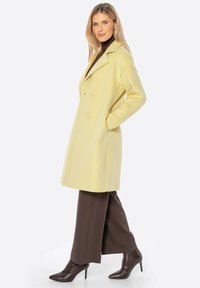 Abrigo amarillo de corte doble con un diseño ajustado. Textura suave, bolsillos laterales, combinado con pantalones de pierna ancha marrones y elegantes botines marrones.