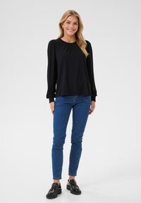 Black long-sleeve top with pleated details, paired with blue slim-fit jeans and black loafers, set against a neutral background.