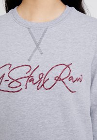 Gros plan d’un sweat-shirt gris à col rond avec un texte rouge brodé en cursive au niveau de la poitrine et une double couture sous le col.