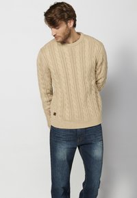 Maglione lavorato a maglia beige con un design a trecce e polsini a coste, abbinato a jeans blu. Il maglione presenta un piccolo logo in pelle.