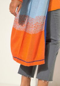 Bufanda de punto en naranja, azul y azul claro con un patrón de degradado. Presenta dobladillo acanalado y una textura suave, colgando de una mano.