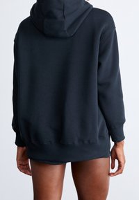 Svart luvtröja med lös passform, ribbade muddar och fåll samt en mjuk textur. Designen inkluderar en diskret halsringning och en söm baktill.