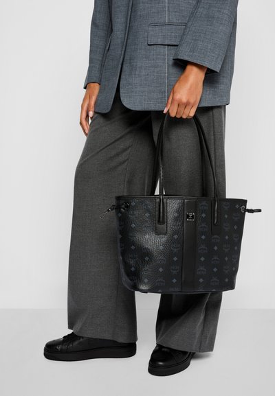 Persona che indossa un completo grigio mentre tiene una borsa a tracolla nera in pelle con un motivo a logo e delle sneaker nere.