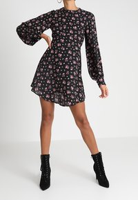 Černé květinové šaty s dlouhými nafouknutými rukávy, přiléhavým živůtkem a rozšířenou sukní. Na šatech se nacházejí malé růžové květy a hladká textura látky. Černé boty na kotníku se šněrováním.