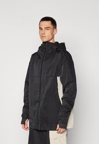 Casaco preto com gola alta, bolsos com fecho éclair e capuz ajustável. Apresenta um painel lateral bege e um tecido texturizado.