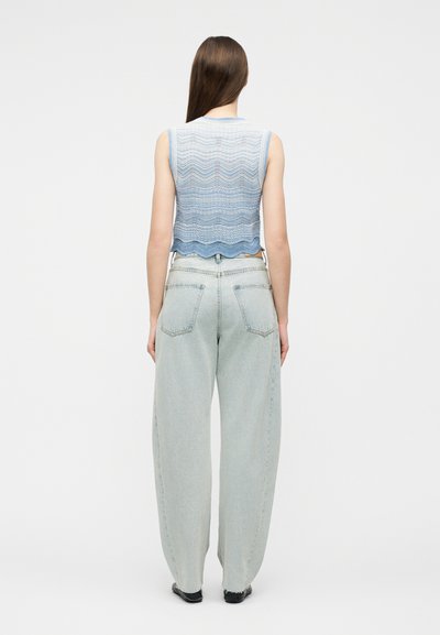 Γυναίκα με μακριά καστανά μαλλιά, φορώντας μπλε ριγωτό πλεκτό τοπ χωρίς μανίκια και φαρδιά τζιν ανοιχτού χρώματος, στέκεται με την πλάτη γυρισμένη.